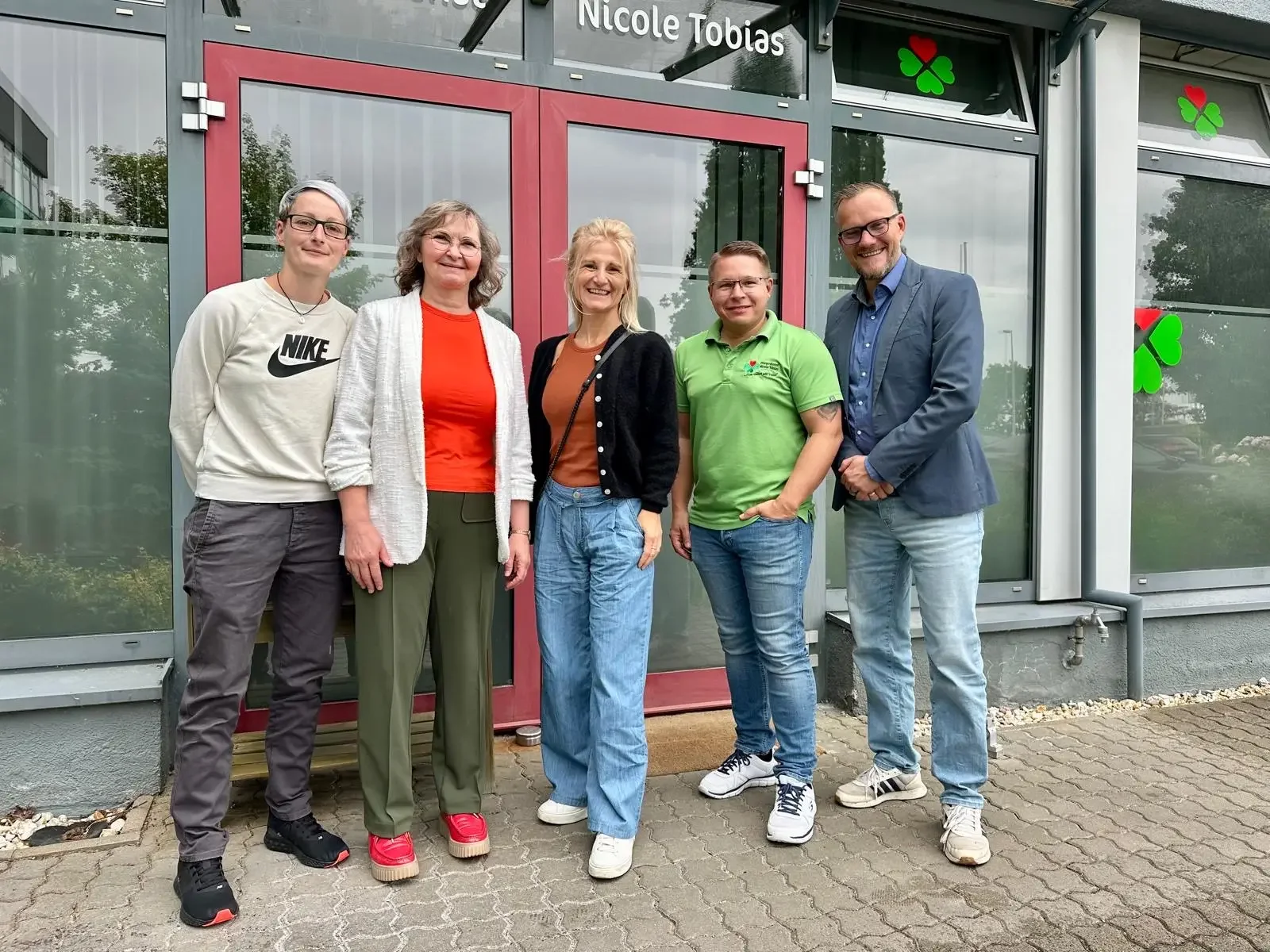 Fünf Personen stehen nebeneinander vor einer Glasfassade mit roter Tür und der Aufschrift „Nicole Tobias“.