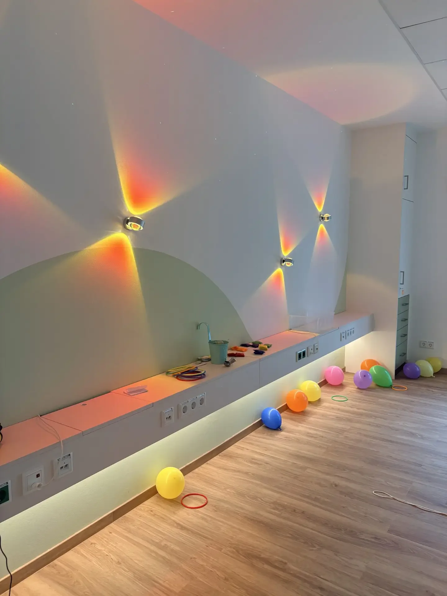 Heller Raum mit langer Sitzbank, Wandleuchten mit bunten Lichtreflexen, Luftballons und Ringen auf dem Holzboden.
