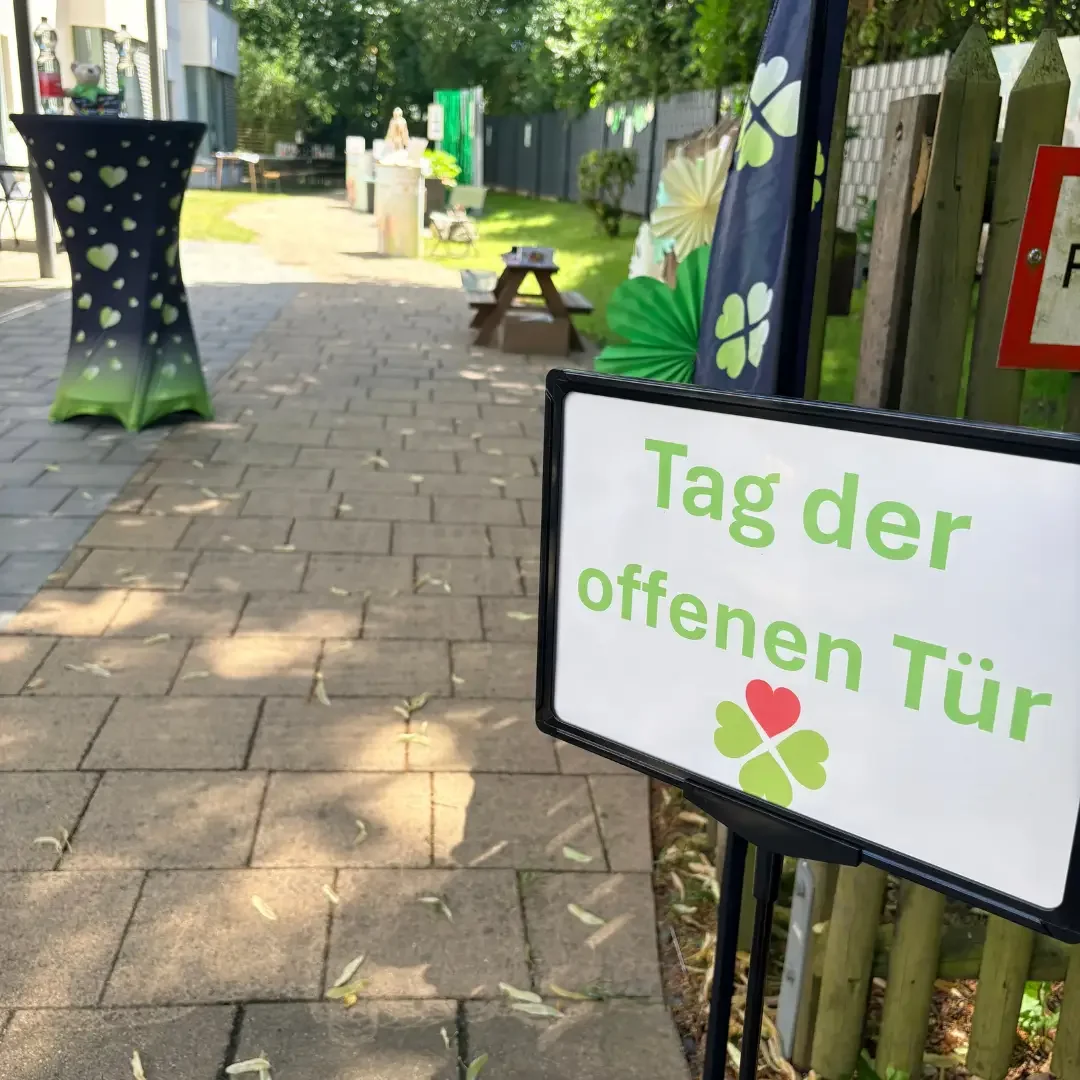 Schild mit der Aufschrift „Tag der offenen Tür“ und Kleeblatt an einem gepflasterten Weg mit Stehtisch im Garten.