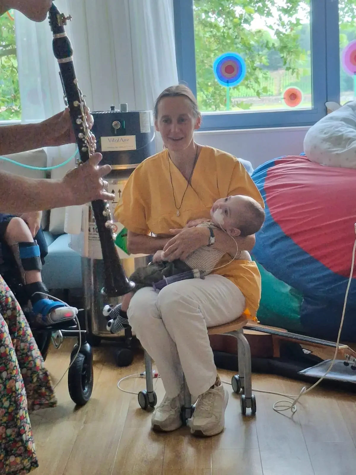 Pflegerin in gelbem Kasack hält ein Kind auf dem Schoß, davor eine Klarinette und ein Gerät mit Aufschrift „VitalAire“.