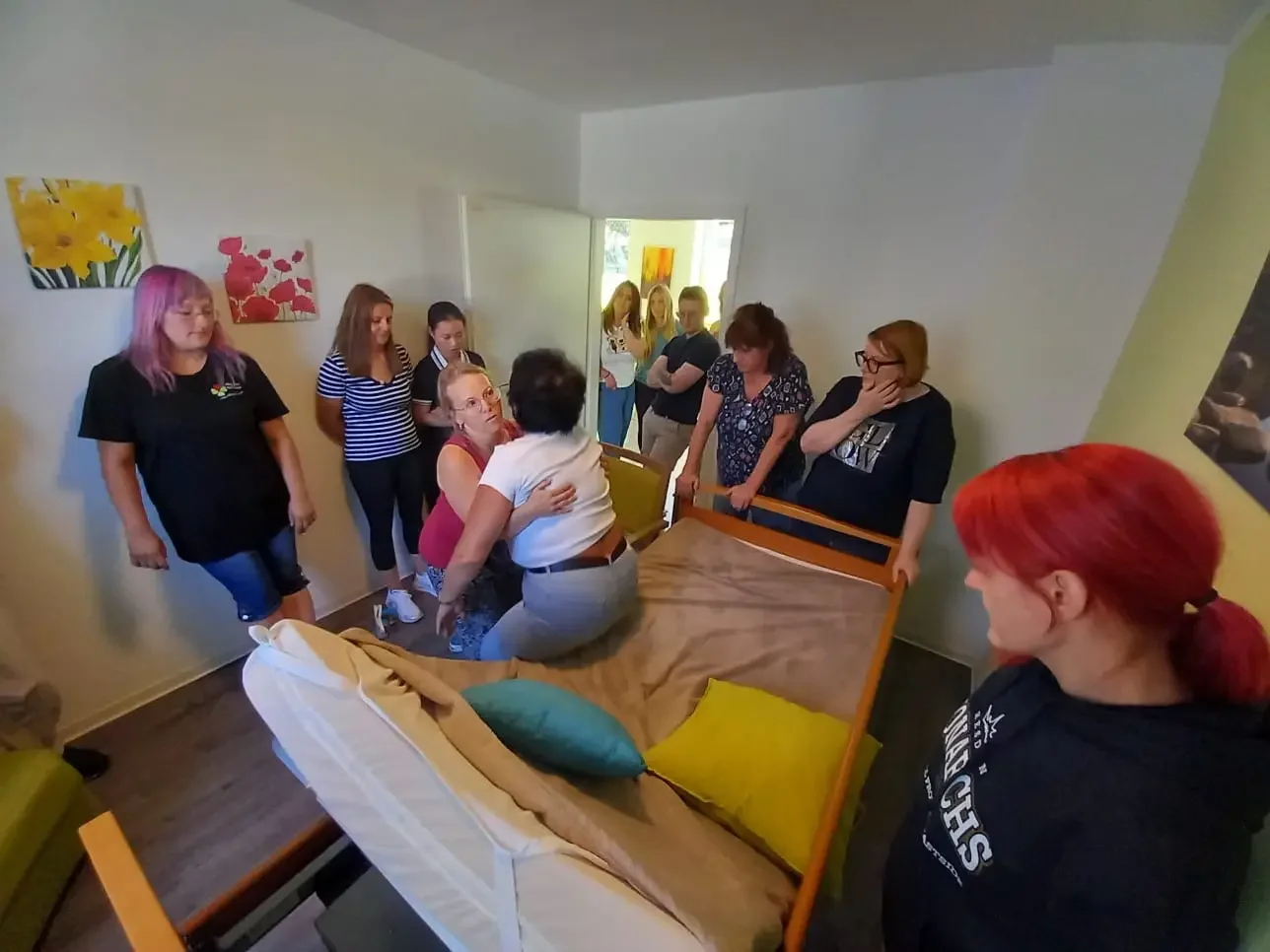 Gruppe von Teilnehmerinnen beobachtet in einem Zimmer eine Transferübung mit einer Person am Pflegebett.