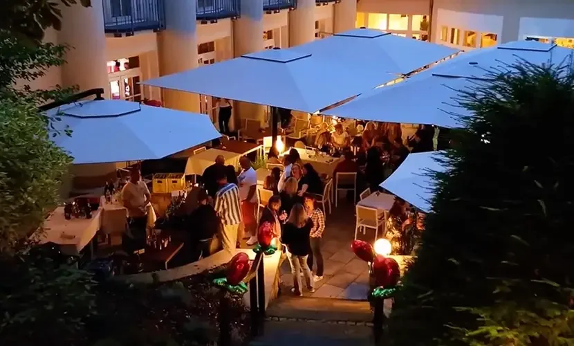 Abendliche Gartenterrasse mit großen weißen Sonnenschirmen, Tischen und vielen stehenden Gästen zwischen Hecken.