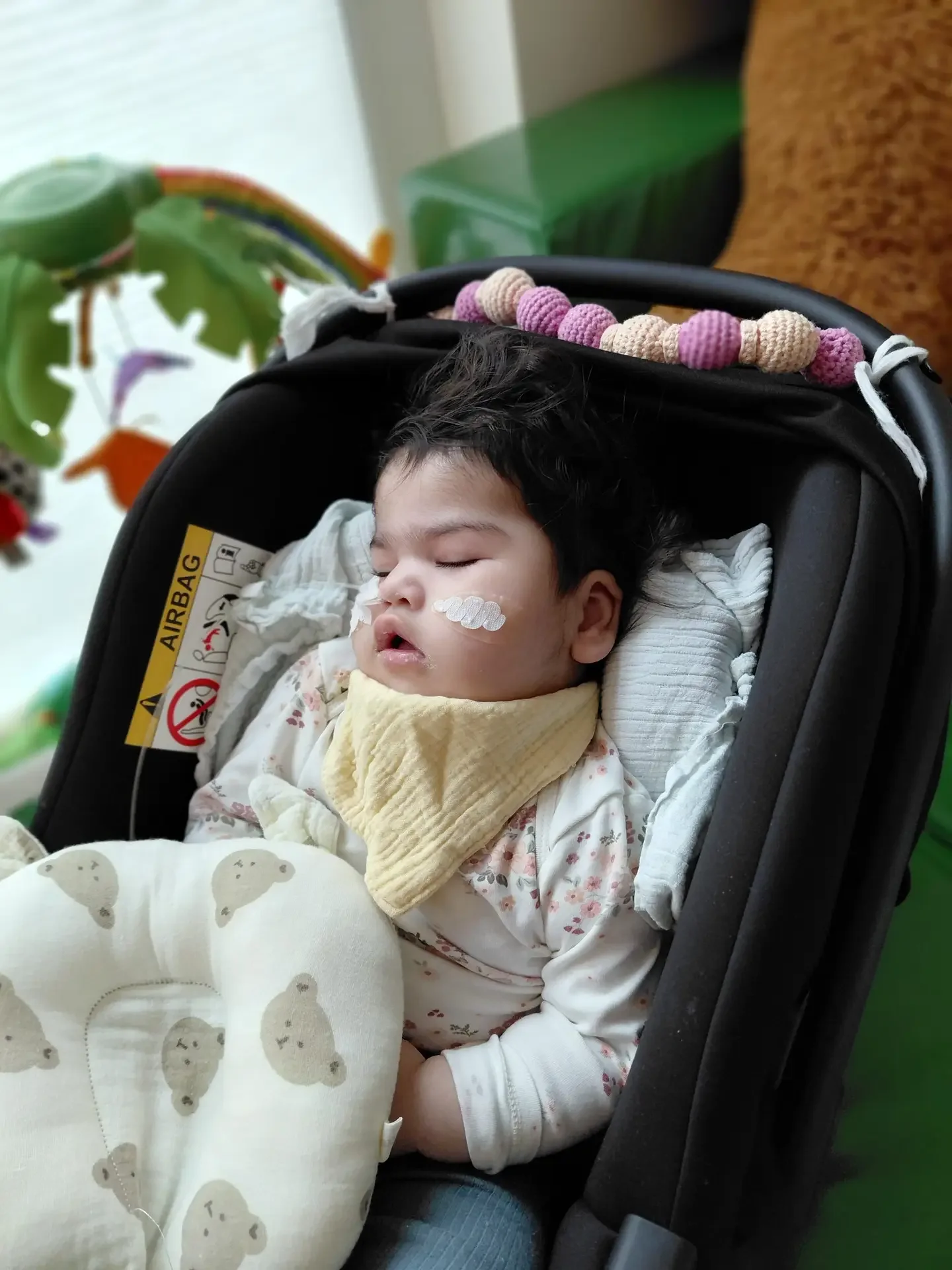 Schlafendes Kleinkind mit Nasensonde und gelbem Halstuch in einer Babyschale mit Aufkleber „AIRBAG“.