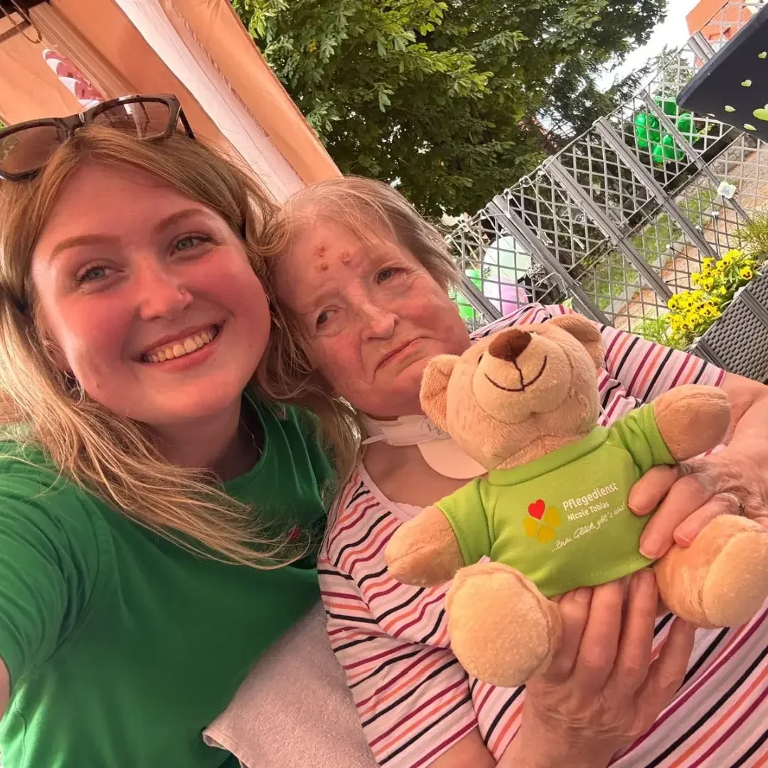 Junge Frau im grünen Shirt und ältere Frau im Liegestuhl halten einen Teddy mit Aufdruck „Pflegedienst Nicole Tobias“.