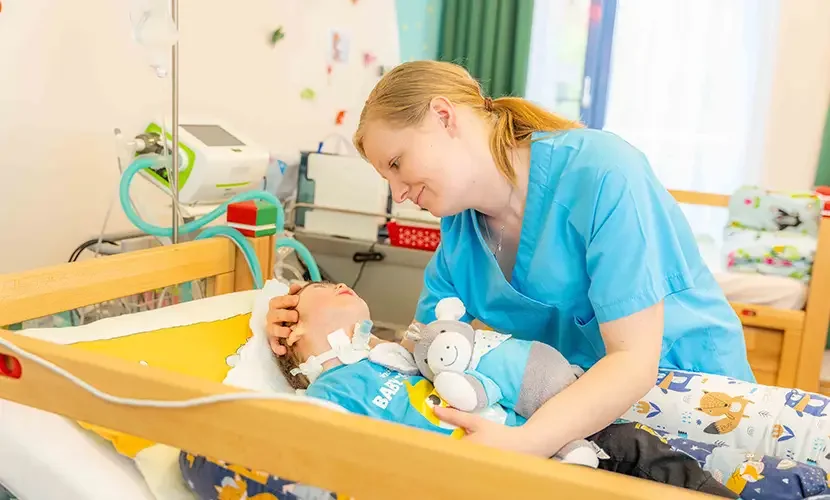 Pflegerin beugt sich lächelnd über ein Kleinkind mit Trachealkanüle im Kinderbett