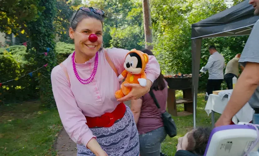 Frau mit roter Clownsnase und pinker Perlenkette hält ein Plüschtier in die Kamera, im Hintergrund ein Gartenfest.