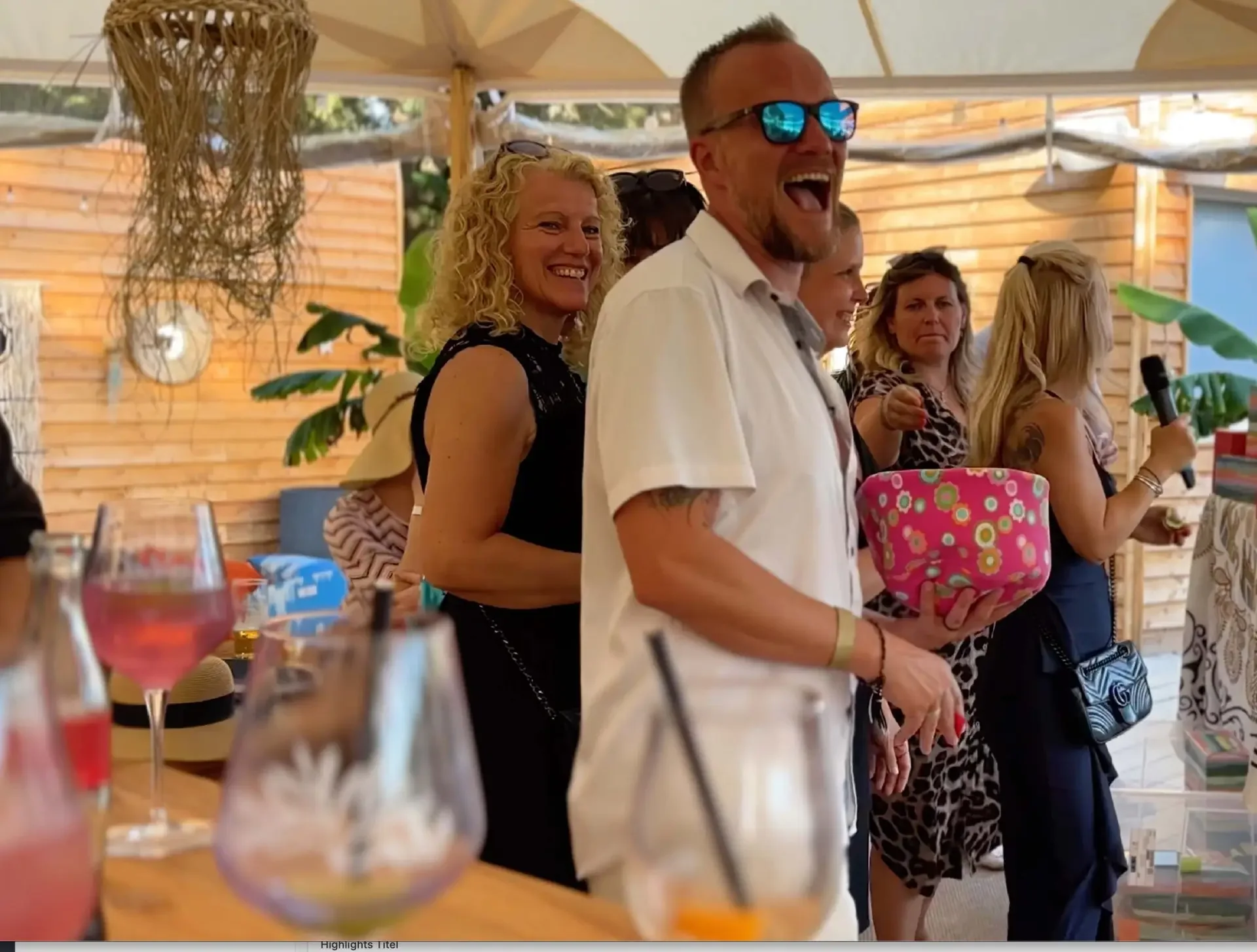 Lachende Gäste unter Sonnenschirmen bei einer Feier im Freien, ein Mann mit Sonnenbrille hält eine geblümte Schüssel.