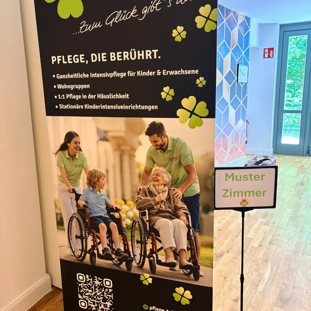 Rollup-Banner „PFLEGE, DIE BERÜHRT.“ mit Pflegekräften und Rollstuhlfahrern neben einem Schild „Muster Zimmer“ im Flur.