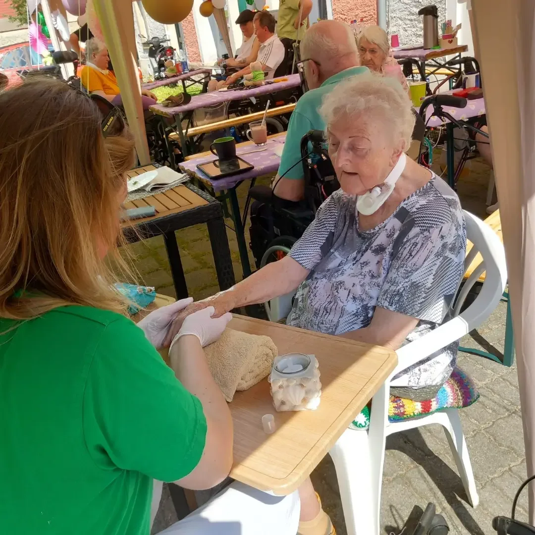 Pflegerin im grünen Shirt mit Handschuhen hält die Hand einer älteren Frau am Tisch bei einem Fest im Freien.