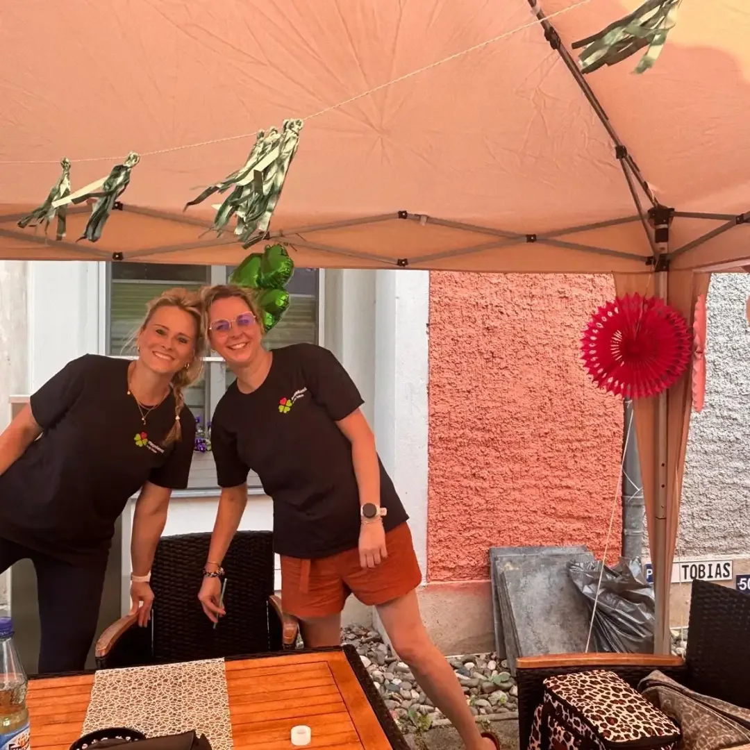 Zwei Frauen in dunklen T-Shirts stehen unter einem Pavillon an einem Holztisch, an der Wand ein Schild „TOBIAS“.