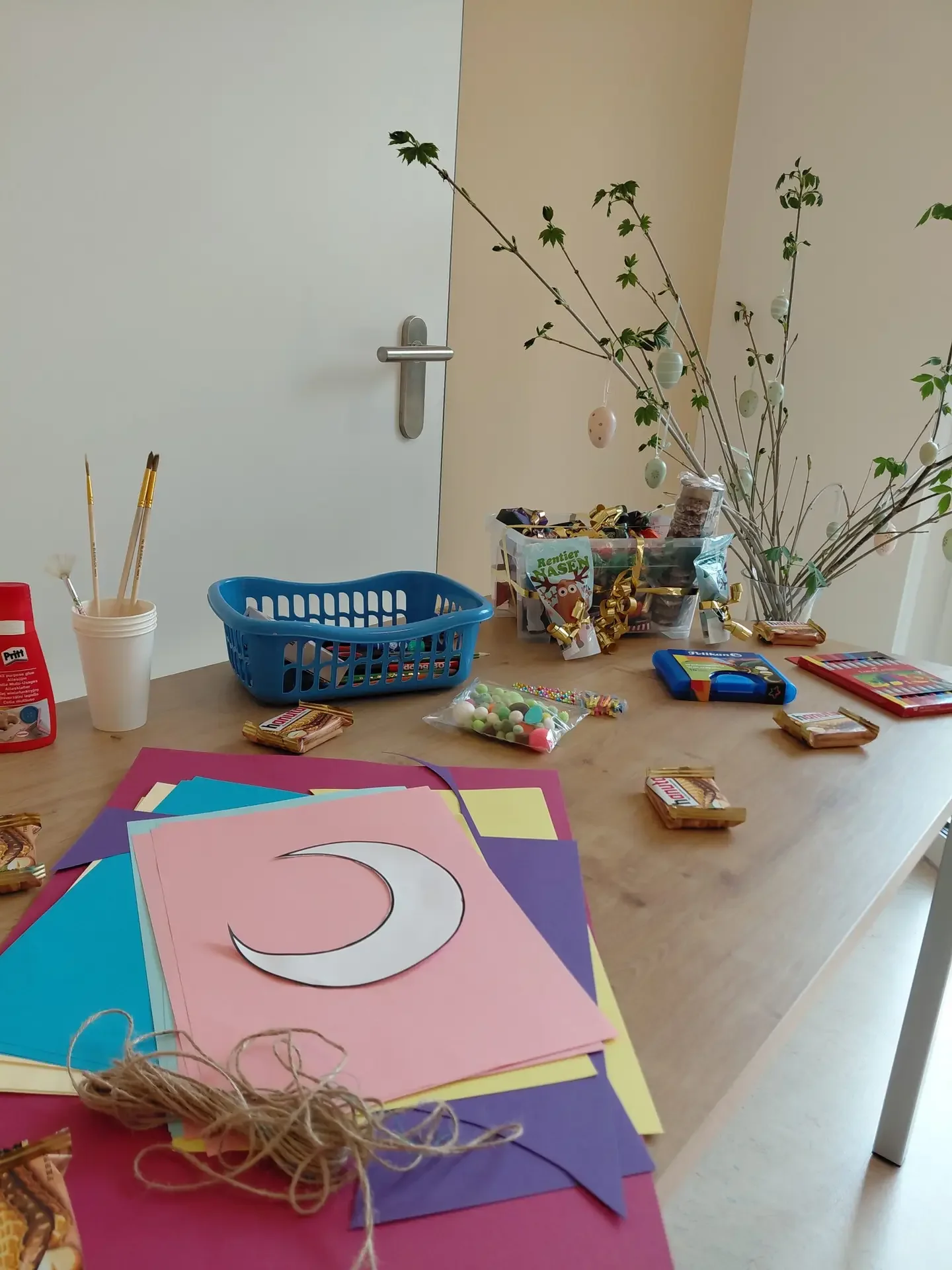 Basteltisch mit buntem Papier, Mond-Ausschnitt, Pinseln, blauem Korb, Süßigkeiten „Nasen“ und Zweigen mit Eiern.