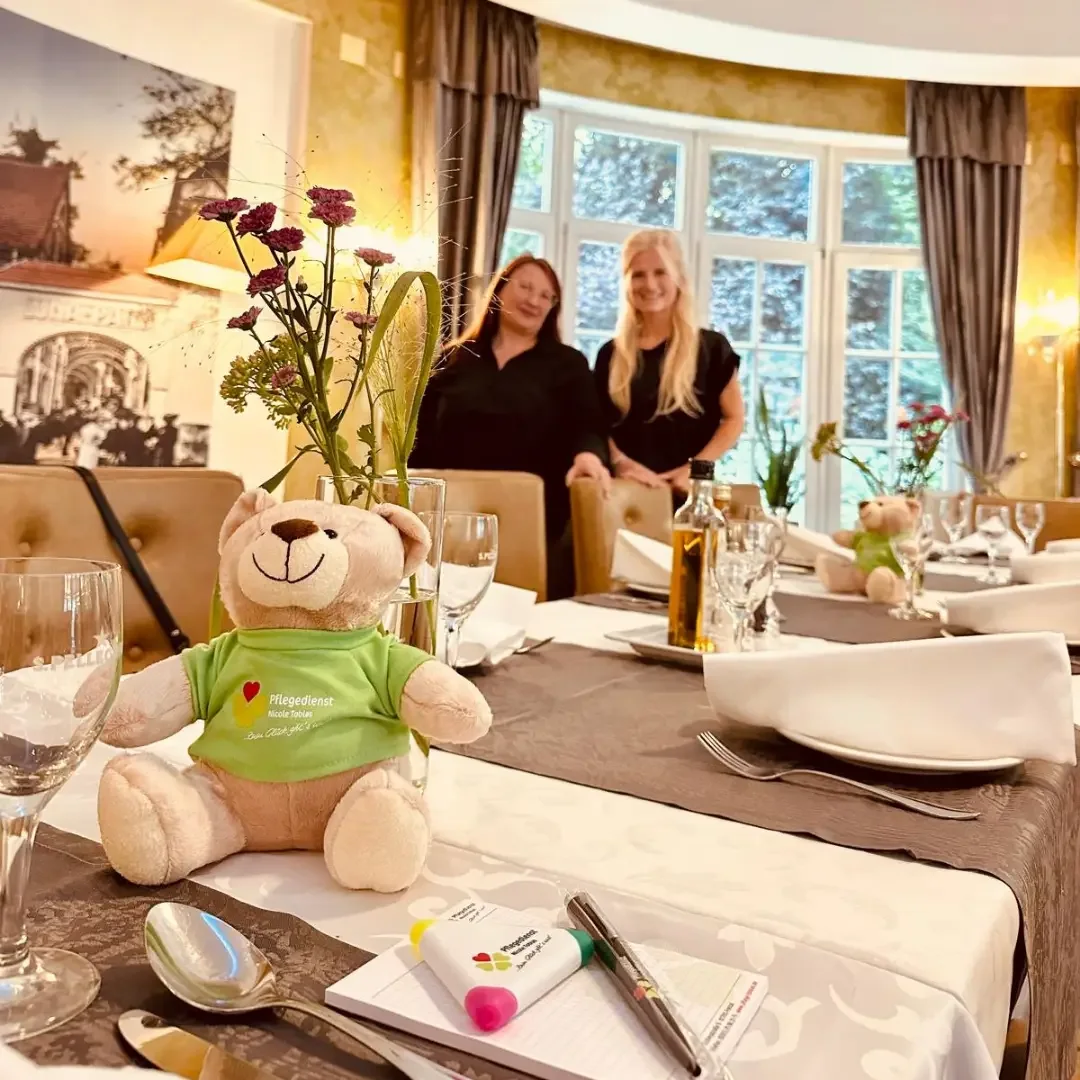 Gedeckter Tisch mit Teddybär im grünen Pflegedienst-Shirt, dahinter zwei Frauen im Restaurant