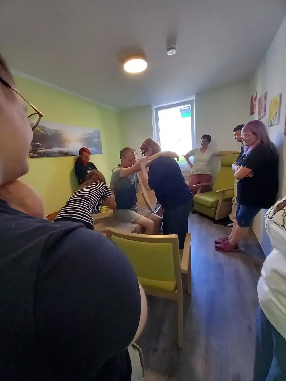 Mehrere Personen üben in einem hellen Raum mit grünen Sesseln das Aufstehen einer sitzenden Person mit Unterstützung.