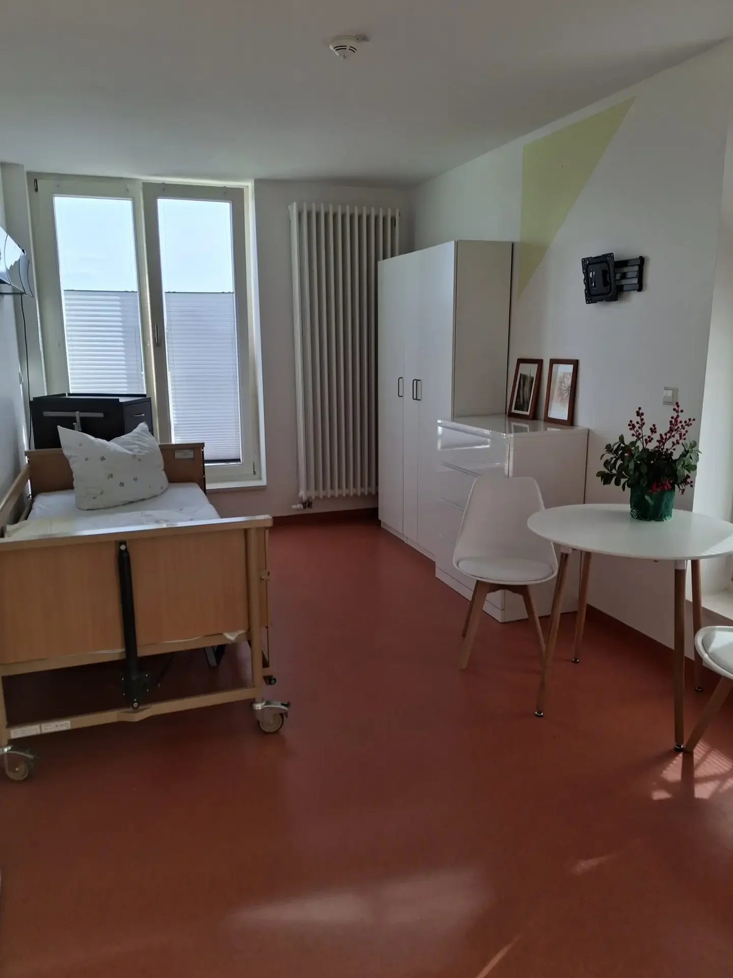 Pflegezimmer mit Pflegebett, weißem Kleiderschrank, rundem Tisch mit zwei Stühlen, Fenster und rotem Bodenbelag.