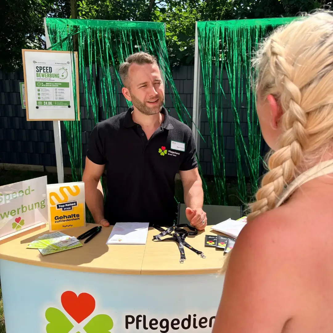 Mann im schwarzen Poloshirt am Messestand „Pflegedienst“ im Gespräch mit blonder Frau, Plakat „Speed-Bewerbung“.