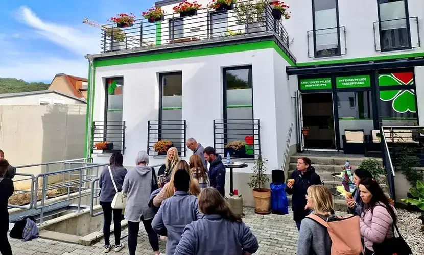 Personengruppe steht vor einem weiß-grünen Gebäude mit Schildern „Ambulante Pflege Wohngruppe“ und „Intensivpflege Tagespflege“.