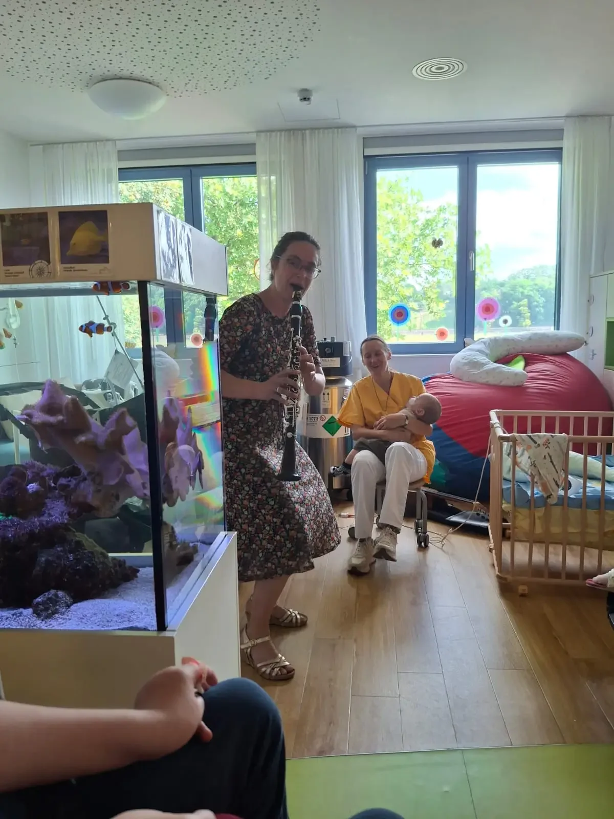 Frau in geblümtem Kleid spielt Klarinette in einem Zimmer mit Aquarium, Gitterbett und Pflegerin mit Kind auf dem Arm.