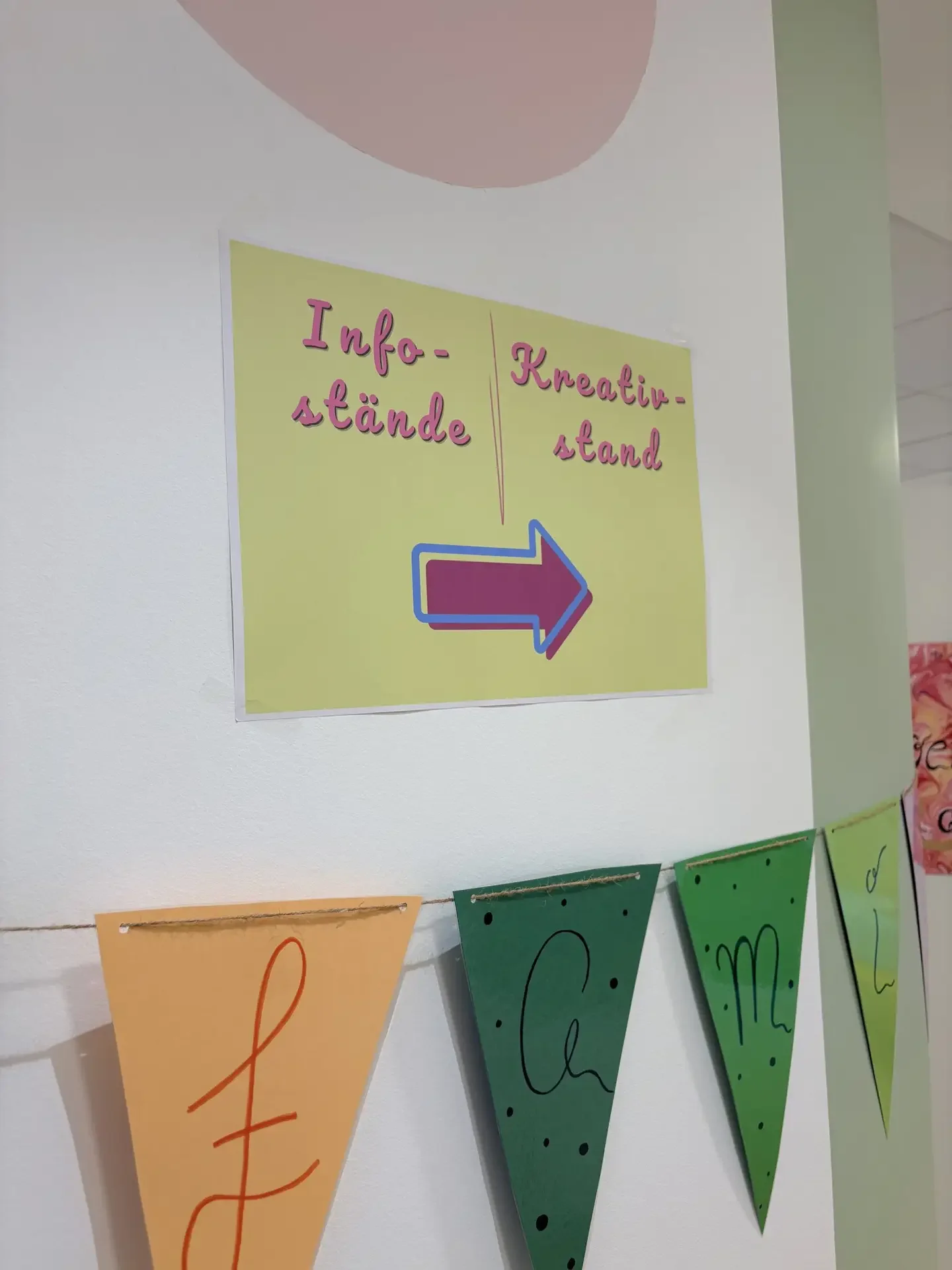 Gelbes Schild an weißer Wand mit pinker Schrift „Info-stände | Kreativ-stand“ und Pfeil, darunter bunte Wimpel.