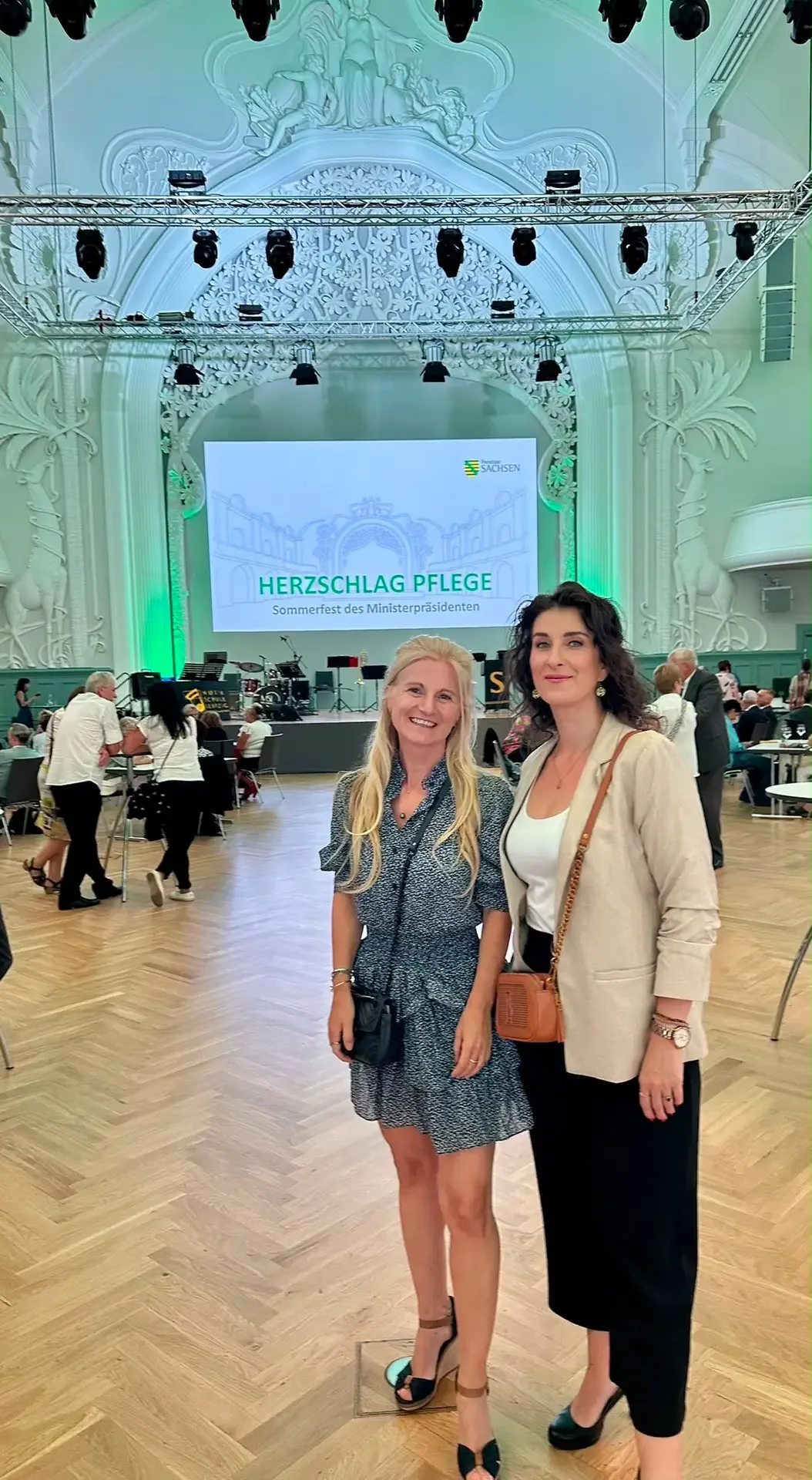 Zwei Mitarbeiterinnen im Festsaal des Sommerfests, dahinter die Leinwand „Herzschlag Pflege“