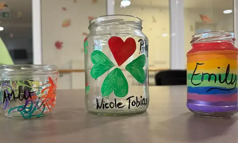 Drei bemalte Einmachgläser auf einem Tisch, beschriftet mit „Nicole Tobias“ und „Emily“, eines mit Kleeblatt und Herz.