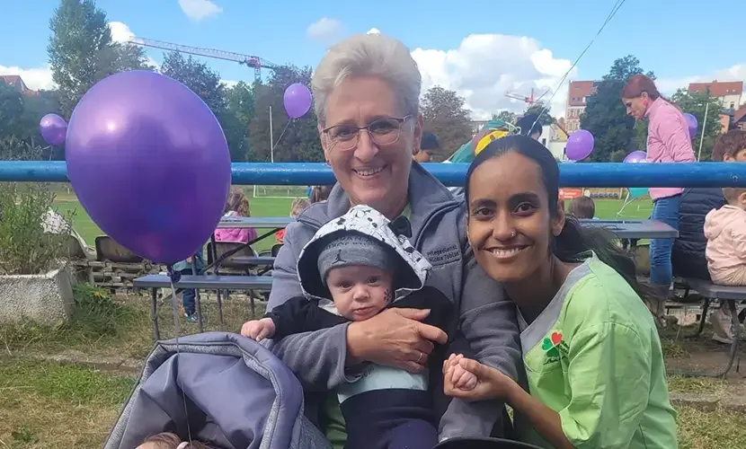 Zwei Frauen mit einem Baby im Arm auf einem Fest im Freien, dahinter Bierbänke und lila Luftballons.