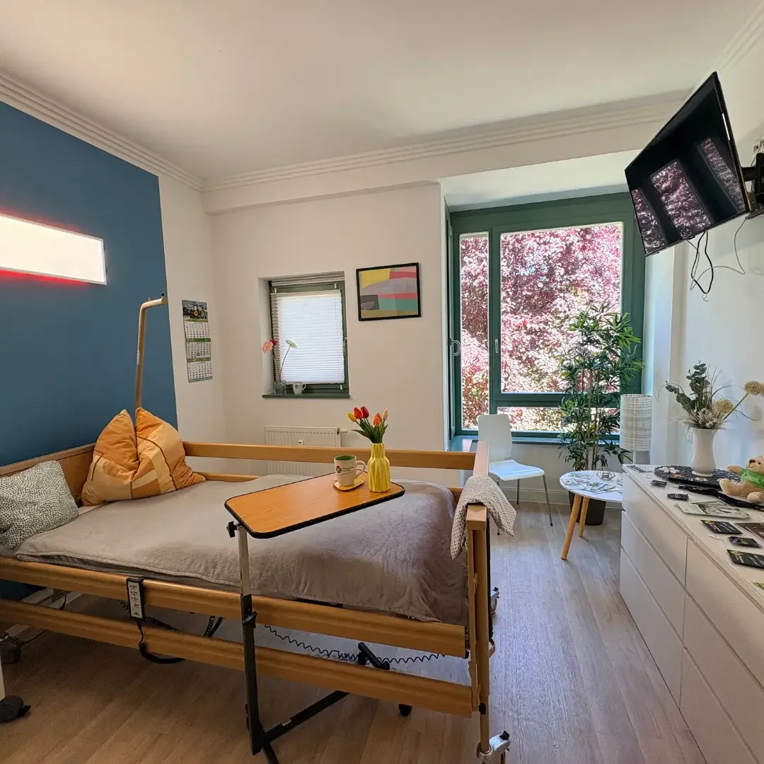 Helles Pflegezimmer mit Pflegebett, Betttisch mit Tulpen, Wandfernseher, blauer Wand und Fenster mit Blick ins Grüne.