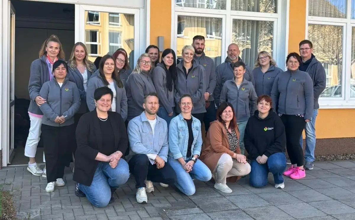 Gruppenfoto von rund 20 Personen in grauen Fleecejacken und Alltagskleidung vor einem Gebäude mit Fensterfront.