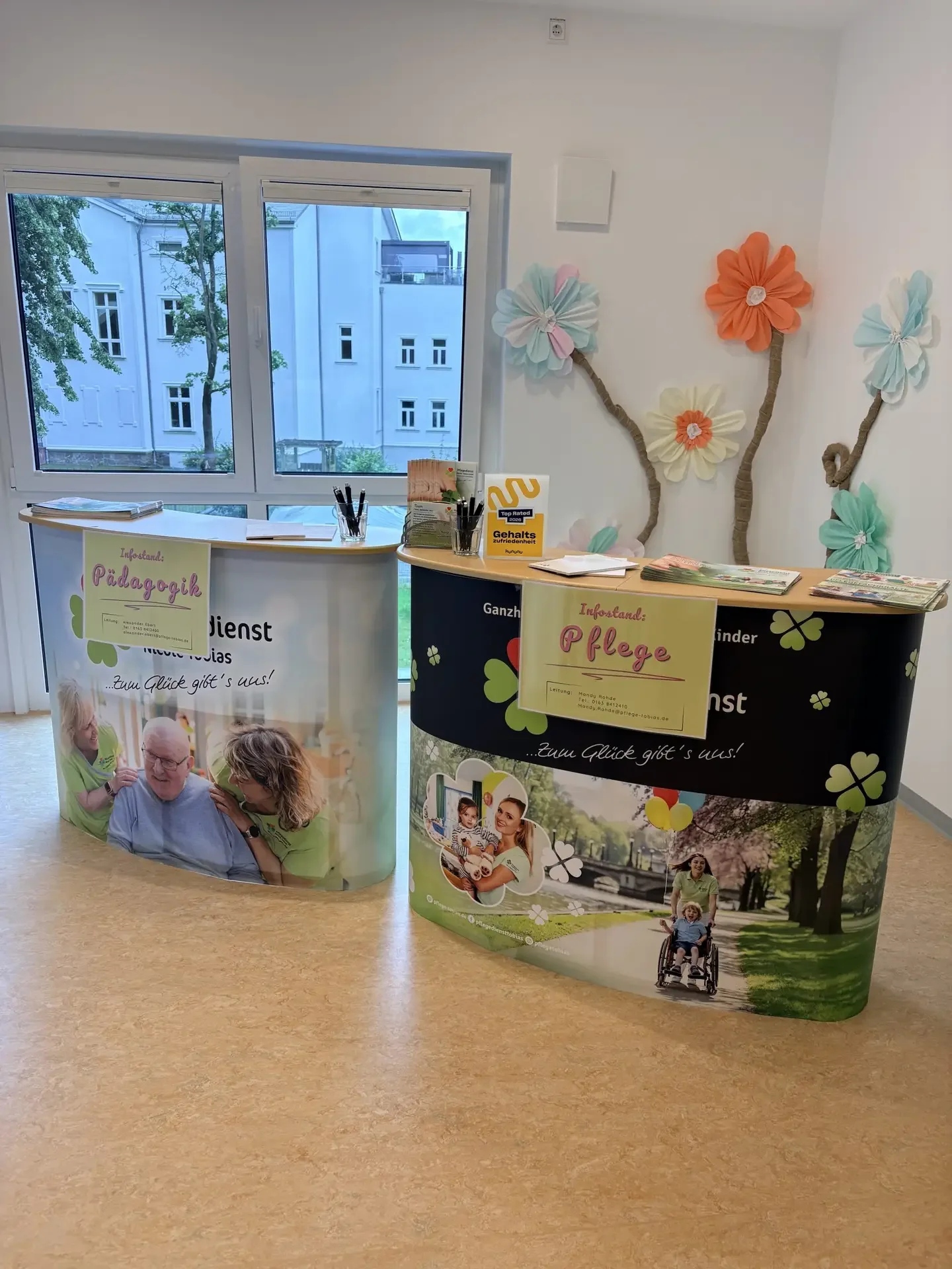 Zwei bedruckte Infotheken mit Schildern „Infostand: Pädagogik“ und „Infostand: Pflege“ vor Papierblumen an der Wand.