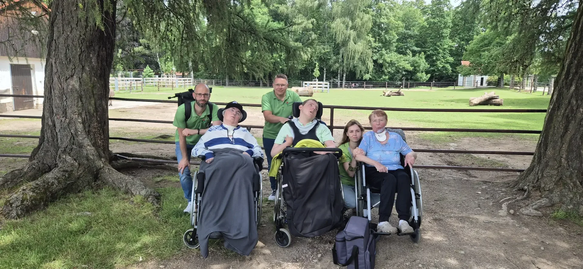 Gruppe mit drei Rollstuhlfahrern und drei Begleitpersonen in grünen Shirts vor einem Zaun mit Weide und Bäumen.