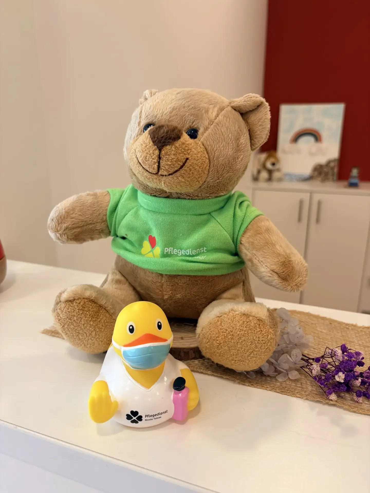 Teddybär im grünen Shirt mit Logo „Pflegedienst“ neben Quietscheente mit Maske und Aufdruck „Pflegedienst Nicole Tobias“.