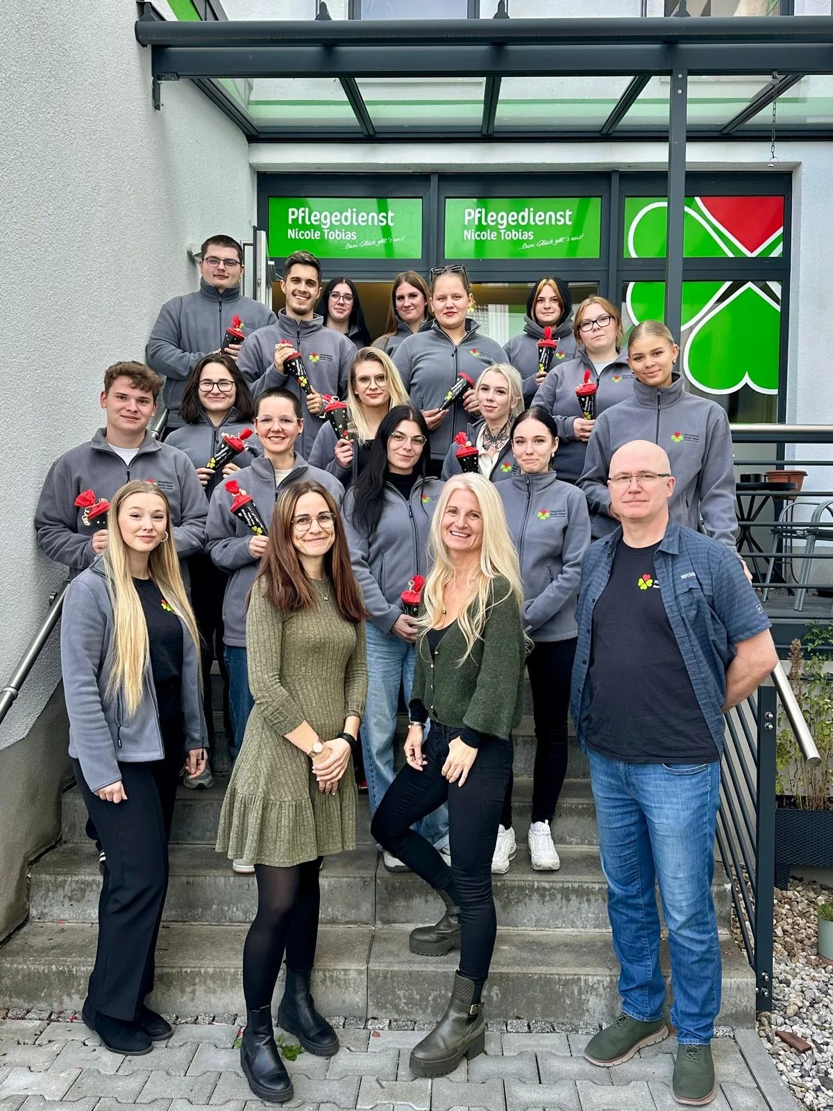 Teamgruppe in grauen Fleecejacken auf einer Treppe vor dem Eingang mit Schriftzug "Pflegedienst Nicole Tobias".
