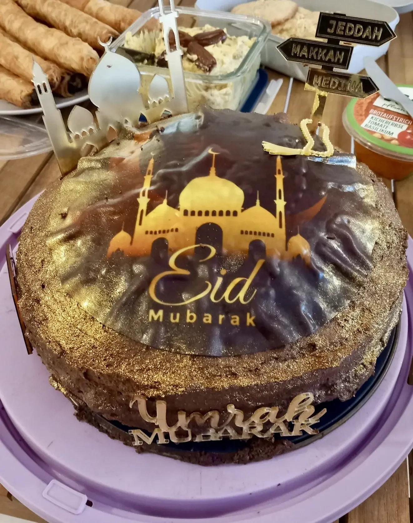 Schokoladentorte mit Aufleger „Eid Mubarak“, Schriftzug „Umrah Mubarak“ und Schildern „JEDDAH“, „MAKKAH“, „MEDINA“.