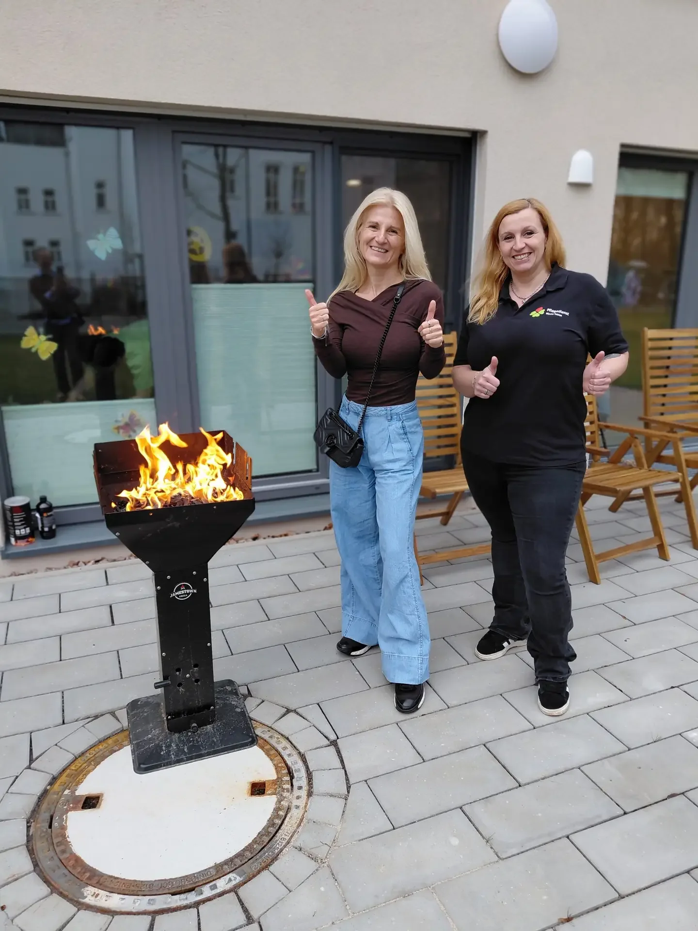 Zwei Frauen zeigen Daumen hoch neben einer brennenden Feuerschale auf einer gepflasterten Terrasse.