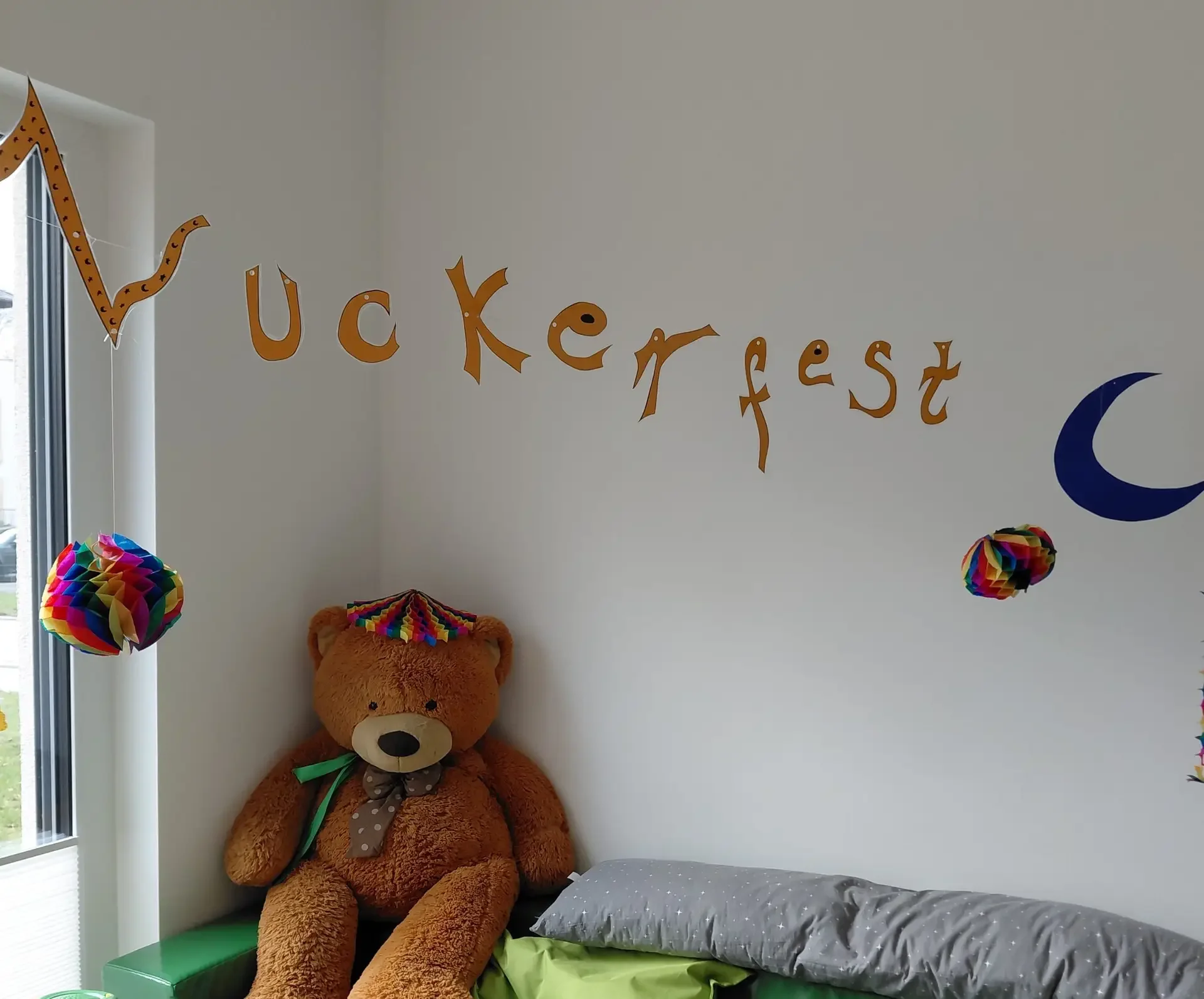 Großer Teddybär mit bunter Krone auf einem Bett unter dem Wandschriftzug „Zuckerfest“, daneben Papierkugeln und Mond.