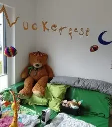 Großer Teddybär auf grüner Couch in einem Zimmer mit Wanddeko „Zuckerfest“, Mond und bunten Lampions.