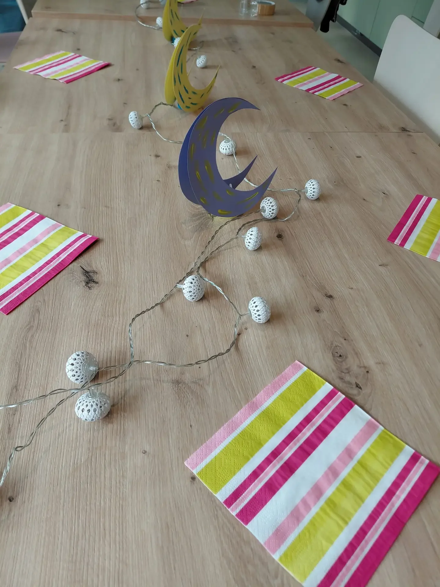 Holztisch mit pink-gelb gestreiften Servietten, weißer Lichterkette und einem blauen Papiermond mit gelbem Stern.