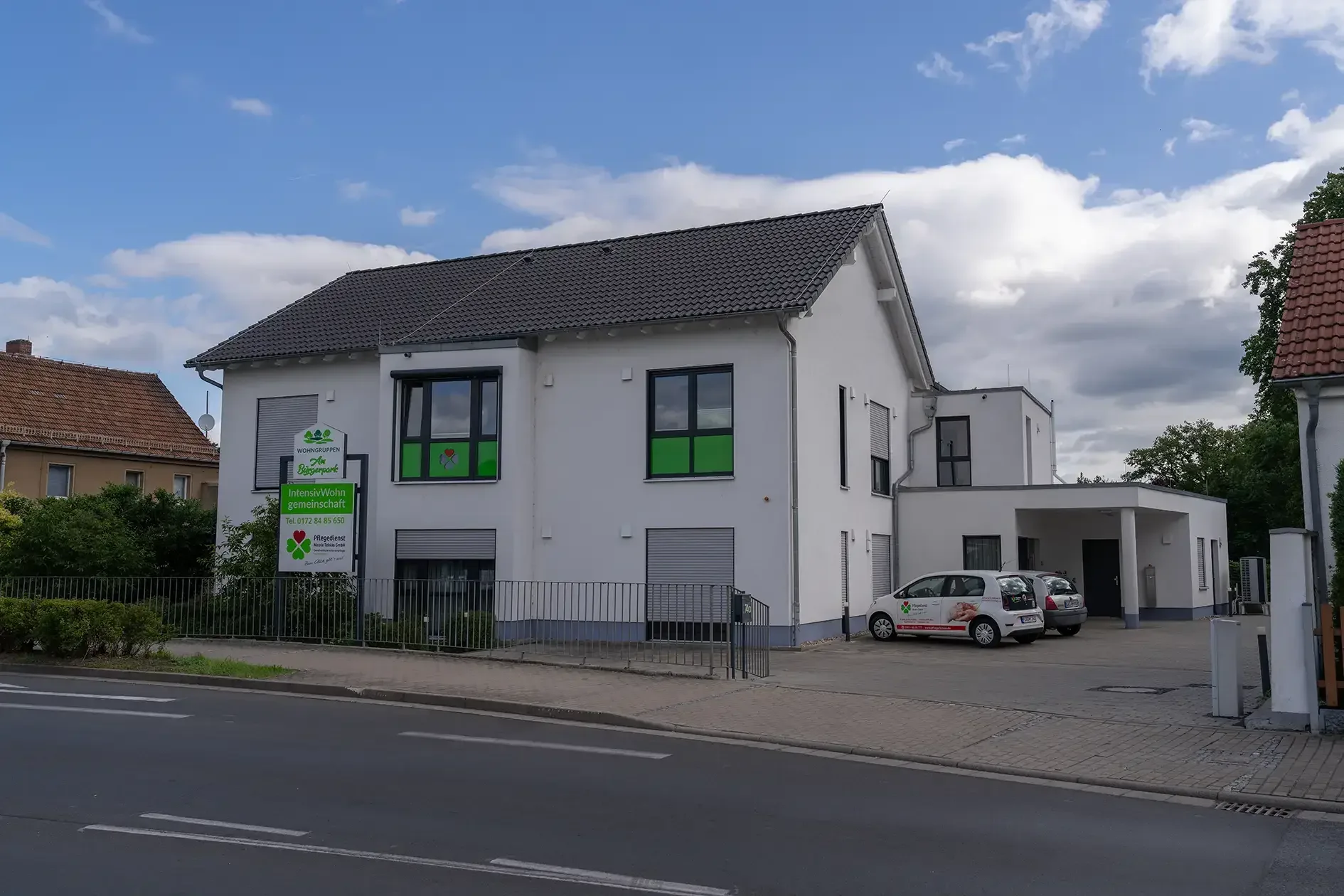 Weißes Wohnhaus mit dunklem Satteldach, Schild „Wohngemeinschaft“ und „Intensiv-Wohngemeinschaft“, davor zwei Kleinwagen.