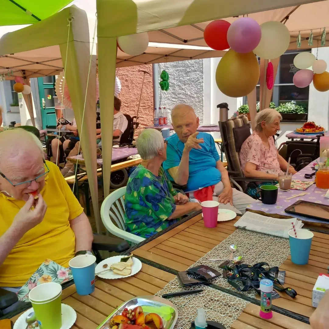 Ältere Menschen sitzen unter Pavillons mit bunten Luftballons an gedeckten Tischen mit Obst, Bechern und Getränken.