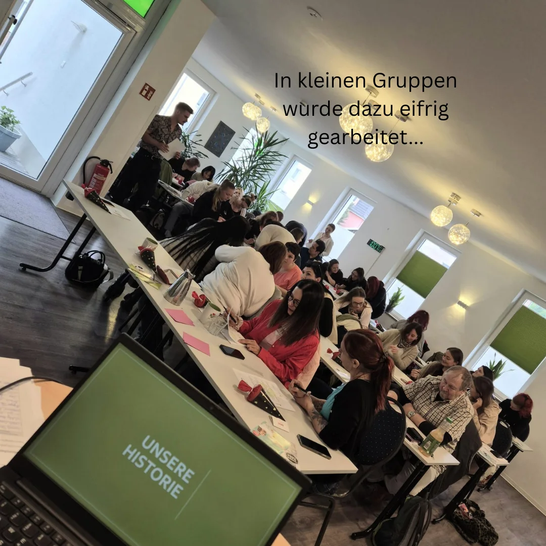 Teilnehmende sitzen in Gruppen an langen Tischen in einem hellen Raum, Laptop zeigt „UNSERE HISTORIE“.