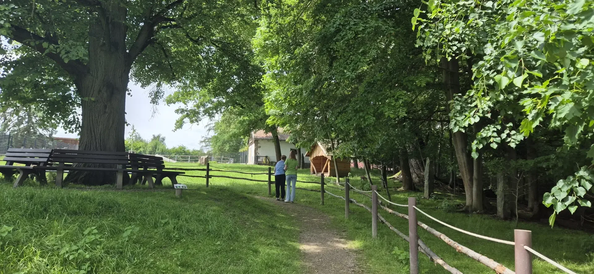 Zwei Personen stehen an einem Holzzaun auf einer Wiese mit Bäumen, Parkbänken und einem Stallgebäude im Hintergrund.