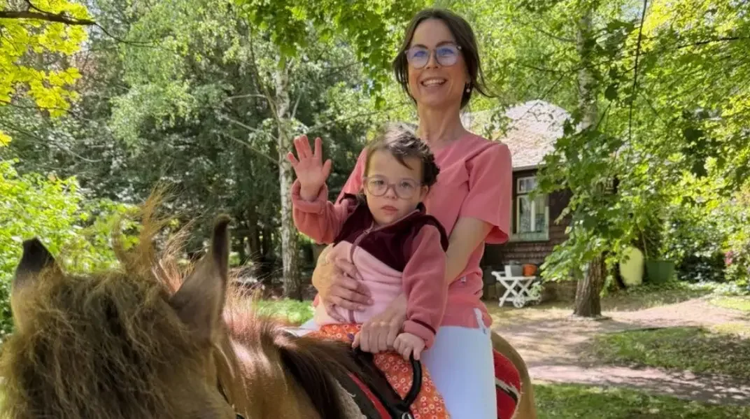 Frau in rosa Shirt sitzt mit einem winkenden Kind mit Brille auf einem Pony in einem sonnigen Garten.