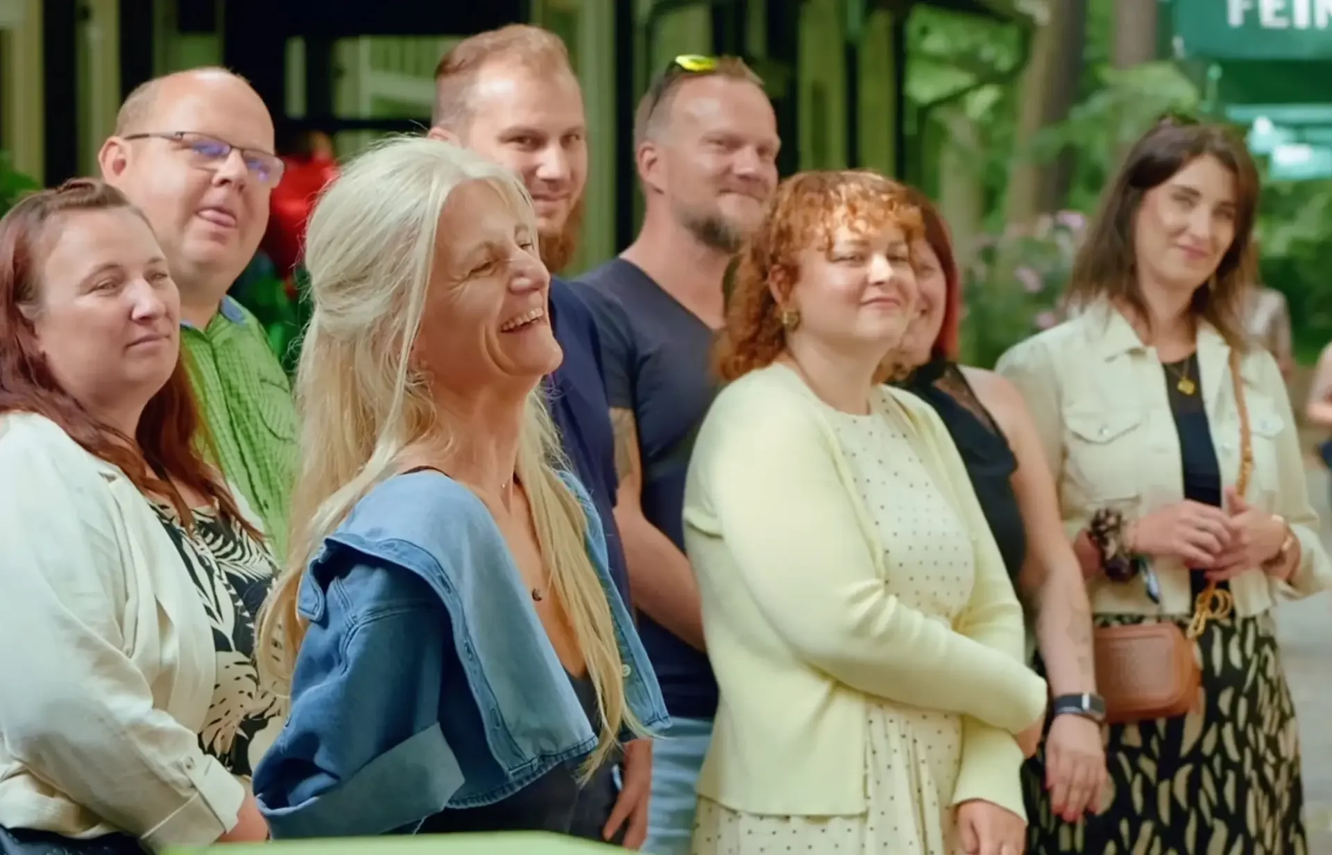 Lächelnde Gruppe von Frauen und Männern steht im Freien beieinander, im Hintergrund ein Schild mit „FEIN“.