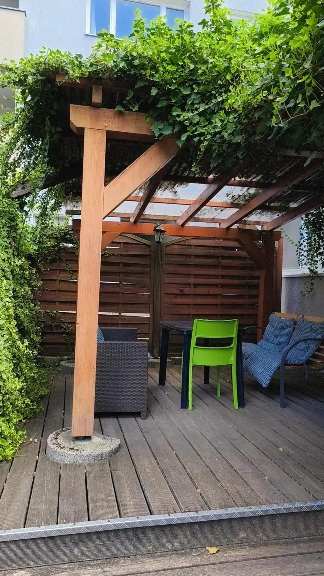 Überdachte Holzterrasse mit efeubewachsener Pergola, grünem Kunststoffstuhl, dunklem Tisch, Rattansessel und Bank mit blauen Kissen.